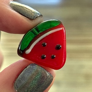 Puede incluir: Un pequeño amuleto de vidrio en forma de sandía con una cáscara verde, semillas blancas y pulpa roja. El amuleto está hecho a mano y tiene un acabado brillante.