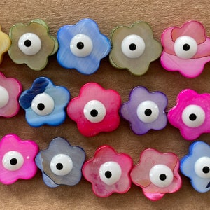 May include: A collection of 14 colorful flower-shaped charms with a single white eye in the center of each charm. The charms are made of a pearlescent material and have a variety of colors, including pink, blue, purple, and green.