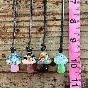 May include: Five glass mushroom pendants on black cords. The mushrooms are all different colors and patterns, including blue and white, black and brown, pink and green, and multi-colored. The pendants are hanging from a wooden surface next to a pink measuring tape.