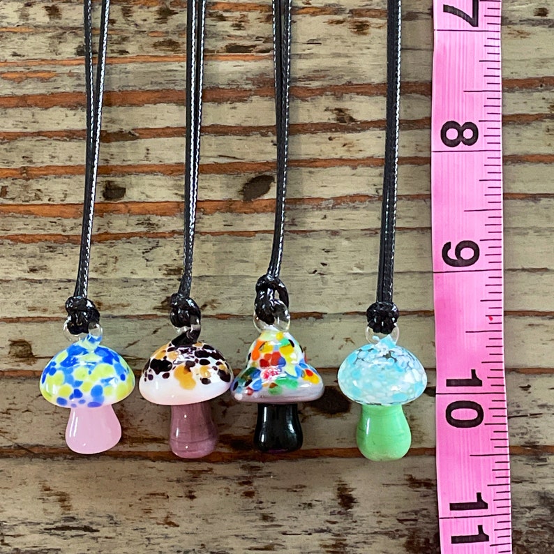 May include: Five glass mushroom pendants on black cords. The mushrooms are all different colors and patterns, including blue and white, black and brown, pink and green, and multi-colored. The pendants are hanging from a wooden surface next to a pink measuring tape.