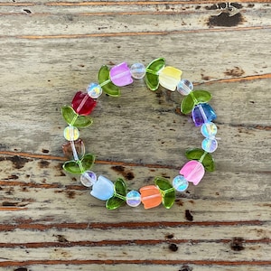 May include: A colorful bracelet featuring tulip-shaped beads in various colors, including red, pink, yellow, and blue. The bracelet also has green leaf accents and iridescent beads, set against a wooden background.