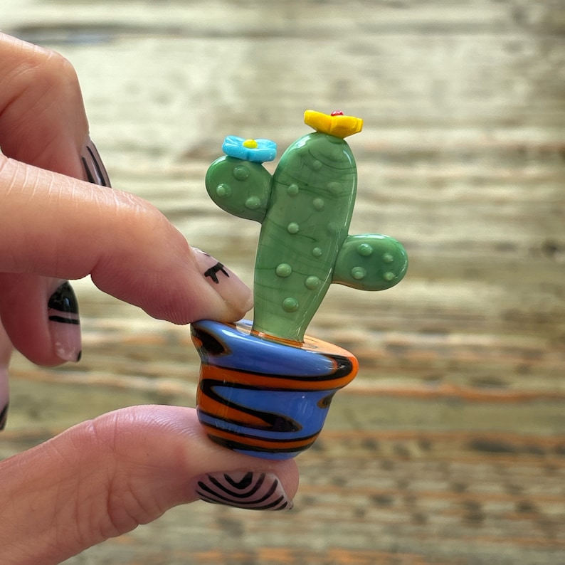 May include: A small green glass cactus with blue and yellow flowers in a blue, orange, and black striped pot.