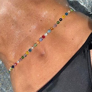 May include: A colorful beaded body chain featuring small, flower-shaped beads in various colors. The chain is worn across the torso, with a black bikini visible in the frame. The beads are arranged in a linear pattern.