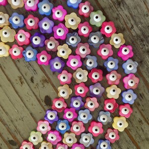 May include: A collection of colorful flower-shaped beads with a single white eye in the center. The beads are made of mother of pearl and are strung together in a curved line.
