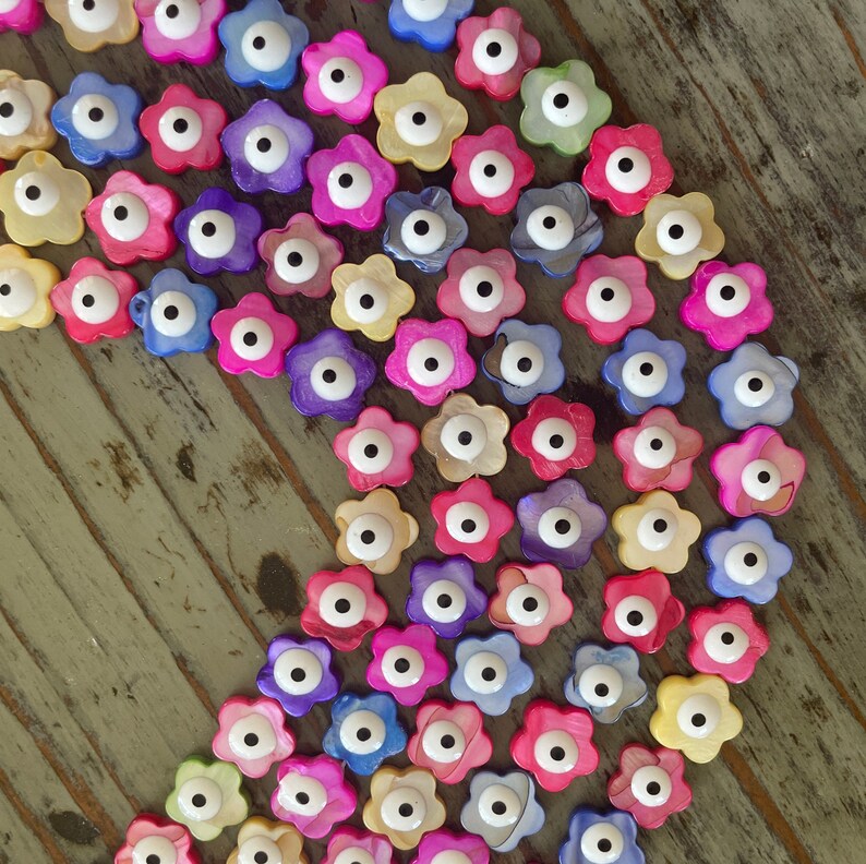 May include: A collection of colorful flower-shaped beads with a single white eye in the center. The beads are made of mother of pearl and are strung together in a curved line.