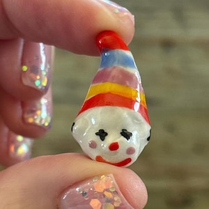 May include: A small, hand-painted clown figurine with a white face, red nose, and a red and rainbow-striped hat. The clown has black eyes and a red smile. The figurine is held between two fingers.