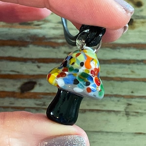 May include: A colorful glass mushroom pendant necklace with a black cord. The mushroom is made of glass with a speckled pattern in shades of blue, green, red, yellow, and white.