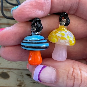 May include: Two glass mushroom pendants on black cords. One mushroom is blue and white with orange stripes, the other is yellow and white with white spots.
