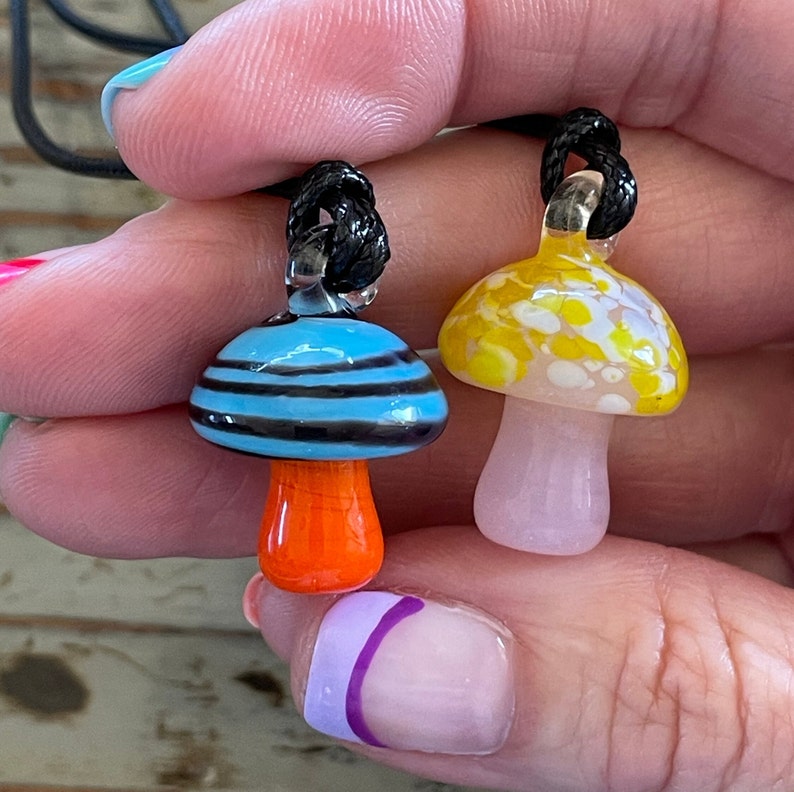 May include: Two glass mushroom pendants on black cords. One mushroom is blue and white with orange stripes, the other is yellow and white with white spots.