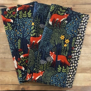 May include: A set of four folded fabric napkins featuring a dark blue background with a repeating pattern of orange foxes, stylized green trees, and yellow flowers. The reverse side of the napkins displays a black and white circular dot pattern.