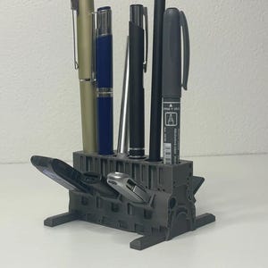 May include: A gray pen holder with a geometric design, holding several pens and a marker. The pens are in various colors, including gold, blue, and black. The marker has the letter "A" printed on it.