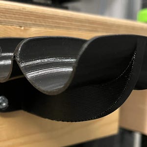 May include: A black, 3D-printed tool holder, mounted on a wooden surface with two screws. The holder features a curved design with three distinct compartments, ideal for organizing tools or small items. The holder is approximately 6 inches long.
