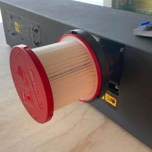 May include: A black rectangular device with a red and white filter attached. The filter has a red cap with white text and a black rim. The device is on a light brown surface.