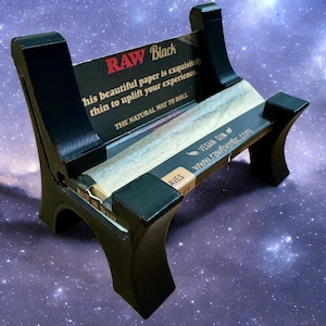 May include: Black rolling paper holder with the text "RAW Black" and "his beautiful paper is exquisitely thin to uplift your experience." The holder is made of black material and holds rolling papers. The text "THE NATURAL WAY TO ROLL" is also visible.