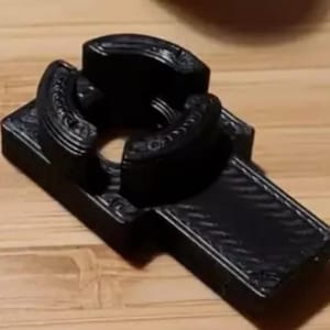 May include: A black, cross-shaped object with a textured surface. The object has a central circular opening and curved, raised elements. The design includes decorative details along the edges, suggesting a functional or decorative item.