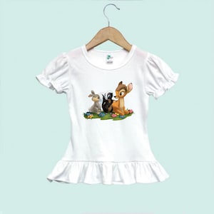 May include: White t-shirt with a ruffle hem featuring a cartoon image of Bambi, Thumper, and Flower from Disney's Bambi.