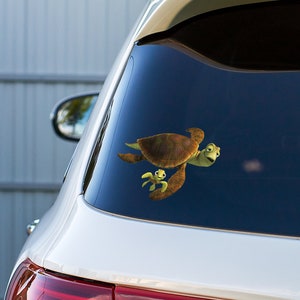 May include: A white car with a sticker of two sea turtles on the back window. The larger turtle is brown with a green head and the smaller turtle is green.