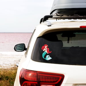 May include: White SUV with a roof rack and a sticker of Ariel the Little Mermaid on the back window. The car is parked in front of a pink lake.