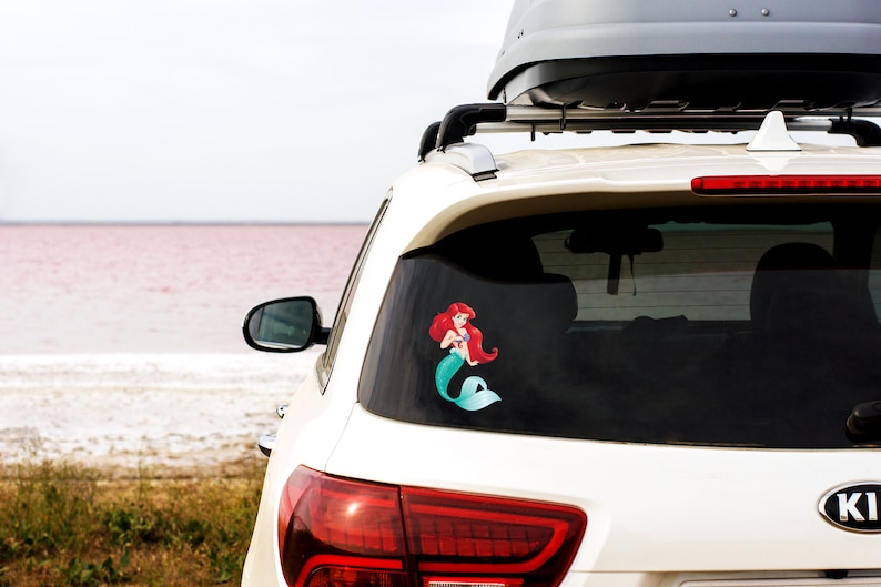 May include: White SUV with a roof rack and a sticker of Ariel the Little Mermaid on the back window. The car is parked in front of a pink lake.