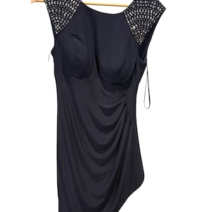 May include: A sleeveless, navy blue dress with sparkling embellishments on the shoulders. The dress features a draped design on the front and a unique asymmetrical hemline. It hangs on a wooden hanger.