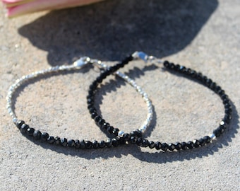 Peut inclure: Deux bracelets délicats en argent avec des perles de pierres précieuses noires. Les bracelets sont posés sur une surface texturée grise.