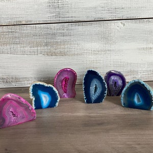May include: Six agate slices in various shades of pink, blue, and purple. The agate slices are polished and have a natural, organic shape. They are arranged in a row on a wooden surface.