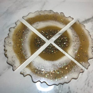 May include: Set of four gold and white resin coasters with a glittery finish. The coasters are shaped like a circle with a wavy edge.