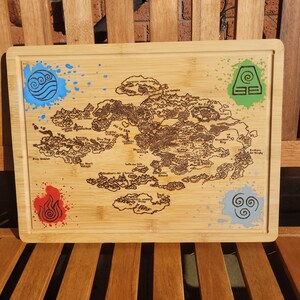 May include: A rectangular bamboo chopping board featuring a map of a fictional world. The map is etched in brown, with colourful paint splatters of blue, red, green, and grey. The board has rounded corners and a smooth surface.