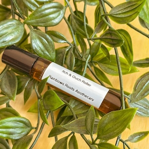 Puede incluir: Un frasco de vidrio marrón con forma de rodillo con una etiqueta blanca que dice "Itch & Ouch Roller" y "Rainbows Roots Apothecary". El frasco está rodeado de hojas verdes.