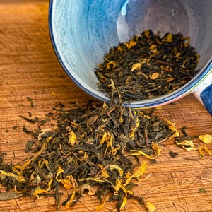 May include: A blue and white ceramic bowl tipped over, spilling loose leaf tea onto a wooden surface. The tea is a mix of green leaves and yellow flower petals. The bowl has a blue interior and a patterned exterior.
