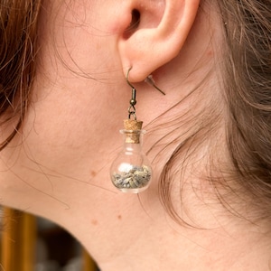 May include: A pair of earrings featuring small glass bottles filled with dried herbs, hanging from gold-toned hooks.