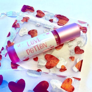 May include: A pink and clear glass bottle with a pink cap and a white label that says "LOVE POTION" in pink lettering. The bottle is sitting in a white mesh bag with a red heart pattern.