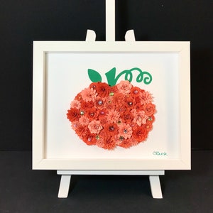 Op de afbeelding: Een ingelijst kunstwerk met een pompoen gemaakt van oranje en rode papieren bloemen, versierd met glinsterende edelstenen. De pompoen heeft een groene stengel en wordt tentoongesteld op een wit schildersezel. De handtekening van de kunstenaar staat in de rechter benedenhoek.