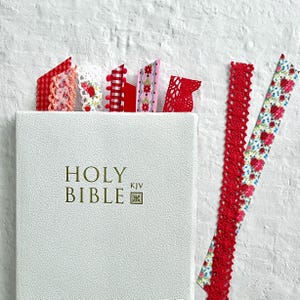 Peut inclure: Une Bible blanche avec une inscription dorée et un marque-page en ruban rouge, blanc et rose. Le marque-page est fait de dentelle et de ruban.