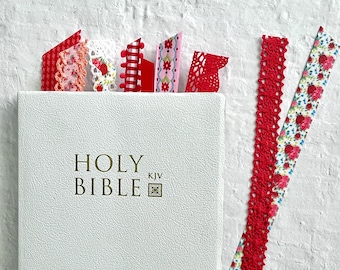 Puede incluir: Una Biblia blanca con una inscripción dorada y un marcapáginas de cinta roja, blanca y rosa. El marcapáginas está hecho de encaje y cinta.