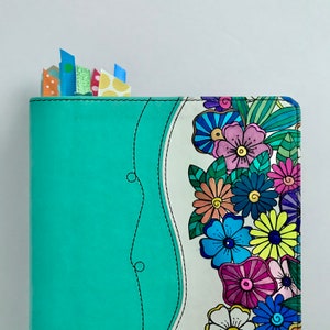 May include: A teal blue notebook with a white cover featuring a colorful floral design. The notebook has a decorative stitch around the edge of the cover.
