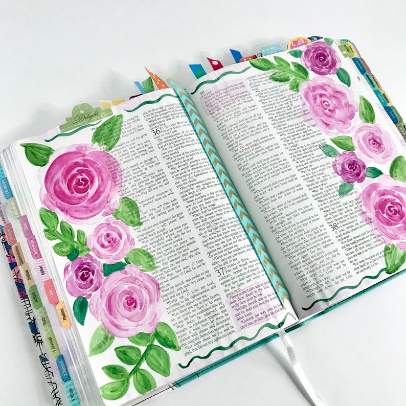 May include: A close-up of an open Bible with watercolor painted pink roses and green leaves on the left page. The right page has text from the book of Psalms, verses 36-38. The pages are decorated with colorful washi tape.