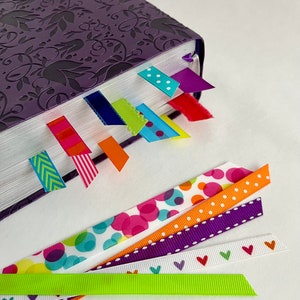 May include: A purple book with a floral design on the cover. The book has several colorful ribbon bookmarks attached to it. The bookmarks are in various patterns, including polka dots, stripes, and arrows.
