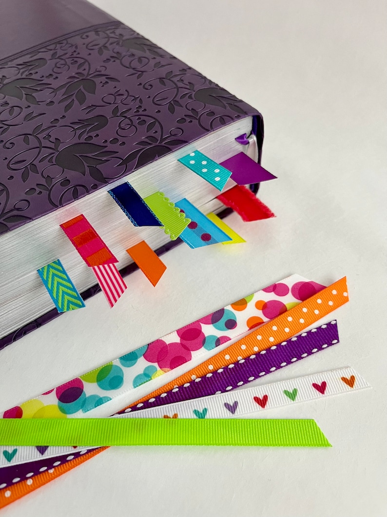 May include: A purple book with a floral design on the cover. The book has several colorful ribbon bookmarks attached to it. The bookmarks are in various patterns, including polka dots, stripes, and arrows.