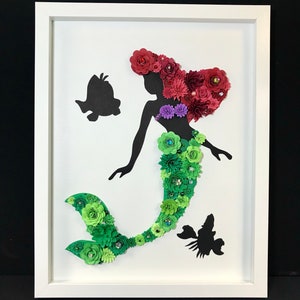 Könnte beinhalten: Gerahmtes Kunstwerk mit einer Meerjungfrau-Silhouette, die mit floralen Elementen verziert ist. Das Haar der Meerjungfrau besteht aus roten Blumen, ihre Flosse ist grün und ihr Oberteil ist lila. Zwei schwarze Fischsilhouetten sind ebenfalls vorhanden.