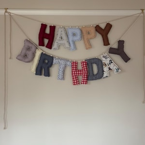 May include: A handmade fabric banner spelling "HAPPY BIRTHDAY". The letters are made from assorted patterned fabrics, including gingham and floral designs. The banner is strung on a neutral-colored cord against a plain wall.