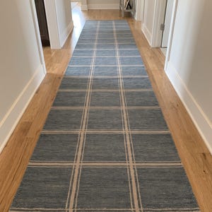 May include: A long, rectangular runner rug featuring a blue base color with a grid pattern formed by thin, light beige lines. The rug is positioned lengthwise on a light-colored wooden floor, extending from the foreground into the distance.