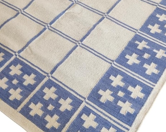 Könnte beinhalten: Ein rechteckiger Teppich mit cremefarbenem Hintergrund und blauem Gittermuster. Der Rand und die Gitterlinien sind blau, mit blau-weißen kreuzförmigen Designs in den Ecken und entlang des Randes. Der Teppich besteht aus gewebtem Material.