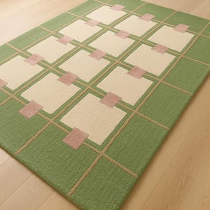 May include: A rectangular rug with a geometric pattern. The rug features a green border and a grid design in tan. Cream-colored squares are interspersed with small pink squares, creating a modern aesthetic.