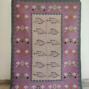 Pode incluir: Um tapete vintage com um padr&atilde;o geom&eacute;trico em tons de roxo, verde e bege. O tapete tem um desenho central de folhas estilizadas e uma borda de formas de diamantes repetidas.