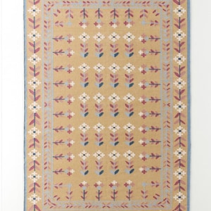 May include: Mustard-colored rectangular rug with a repeating floral pattern in white, pink, and blue. The rug has a blue border and fringe on the short ends. The floral design is symmetrical and covers the majority of the rug's surface.