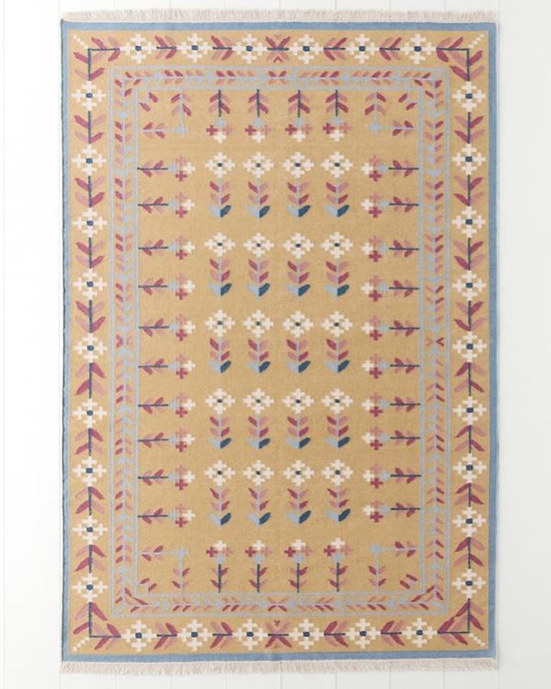 May include: Mustard-colored rectangular rug with a repeating floral pattern in white, pink, and blue. The rug has a blue border and fringe on the short ends. The floral design is symmetrical and covers the majority of the rug's surface.