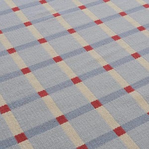 May include: A woven rug with a repeating pattern of light blue, cream, and red squares. The design features diagonal stripes of light blue and cream, intersected by red squares. The rug has a textured appearance.