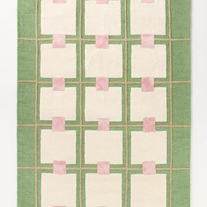 May include: A rectangular rug with a green background and a grid pattern. The grid is made of tan lines, with off-white squares and pink rectangles. The rug has a soft, textured appearance.