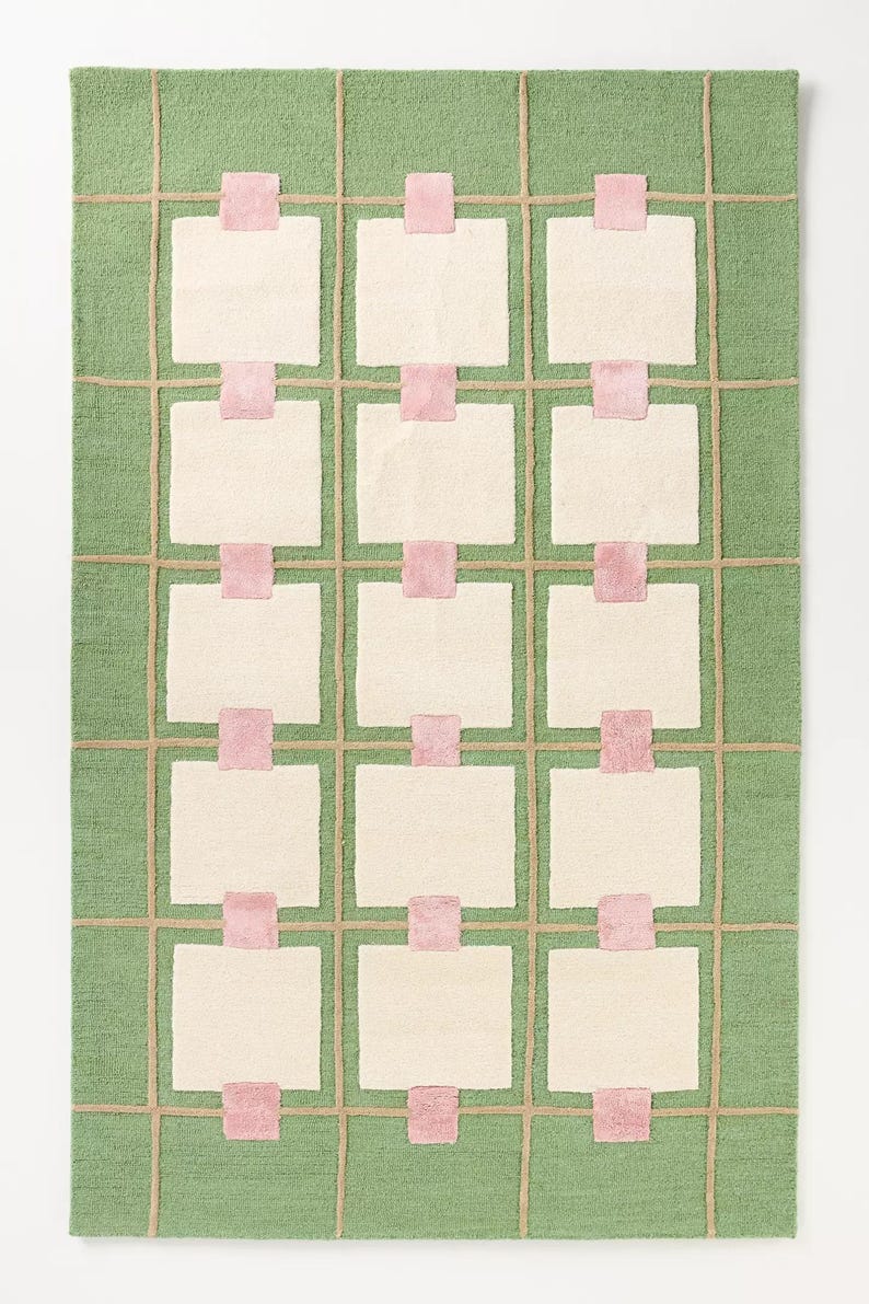 May include: A rectangular rug with a green background and a grid pattern. The grid is made of tan lines, with off-white squares and pink rectangles. The rug has a soft, textured appearance.
